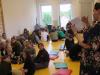 Na zdjęciu grupka dzieci uczestniczących w zajęciach, siedzących na poduszkach i pani bibliotekarka tłumacząca dzieciom zasady zabawy. 