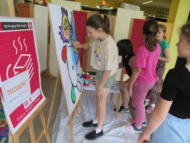Na zdjęciu grupka dzieci stojących przy sztaludze i malujących na niej farbami. 