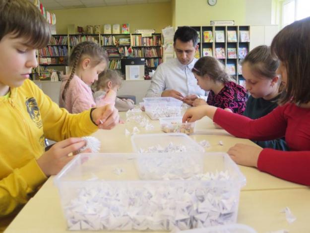 Na zdjęciu grupka dzieci wraz panem, siedzą przy stolikach i wykonują prace origami.