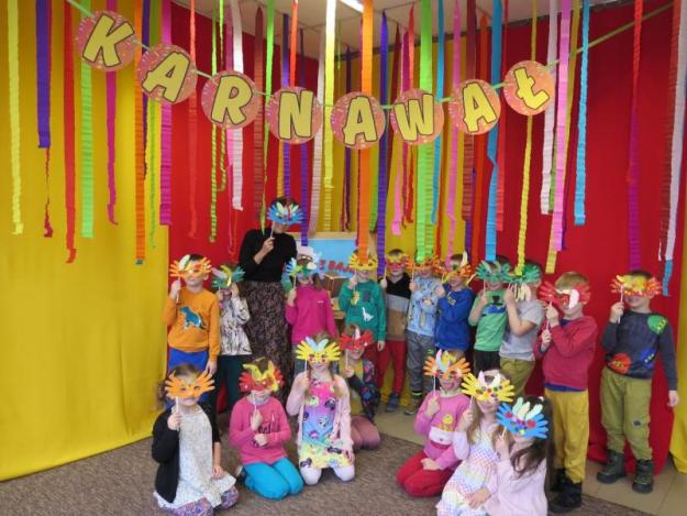 Na zdjęciu dzieci z bibliotekarką w maskach, na tle kolorowej dekoracji.