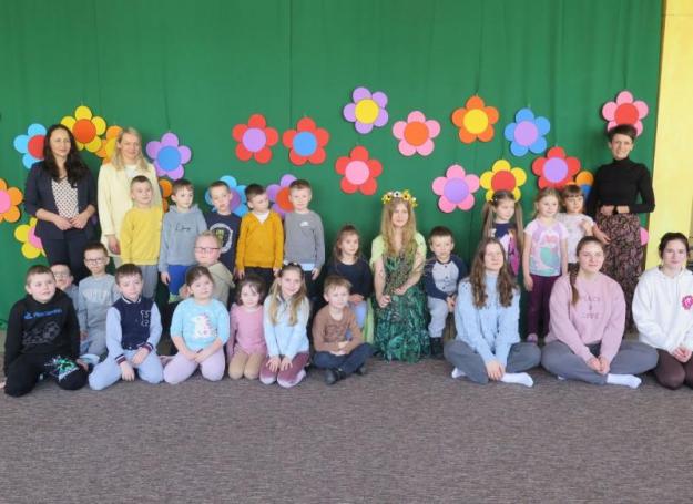 Na zdjęciu spora grupka przedszkolaków wraz z paniami. Zdjęcie wykonane na tle z kwiatami.